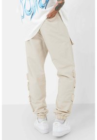 Beżowe spodnie cargo z elastycznymi mankietami, wyposażone w boczne kieszenie i regulowane paski po bokach, zestawione z białymi sneakersami.