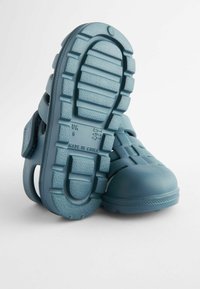 Blå gummiskor för barn med mönstrad, texturerad sula, justerbart spänne och ventilationshål, utformade för komfort och hållbarhet.
