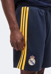 Shorts bleu foncé avec des bandes latérales jaunes et un logo du Real Madrid, portés par une personne avec une main détendue à côté du short.