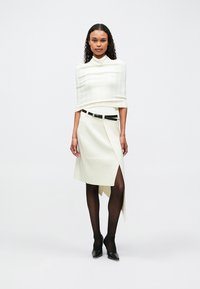 Donna che indossa un top bianco a collo alto e una gonna bianca asimmetrica con spacco, cintola nera, collant trasparenti neri e tacchi neri.