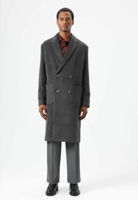 Dunkelgrauer Doppelreihiger Mantel aus Wollmischung mit aufgeschlagenem Revers, zwei Fronttaschen und lockerem Schnitt, ergänzt durch graue Hose und schwarze Schuhe.