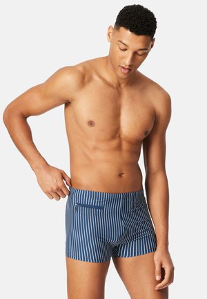 Shirtless man adjusting blue and white vertical striped swim shorts with zippered pocket, looking down, against plain background.