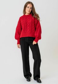 Sarkana adītā džemperis ar kabelīšu rakstiem, augsts kakla izmērs un balloona piedurknes, apvienots ar melnām piegriezuma biksēm un spīdīgām melnām kurpēm.