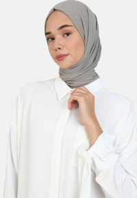 Hijab grigio chiaro in tessuto morbido che si drappeggia attorno alla testa, abbinato a una camicia bianca oversize con una tasca sul petto e maniche arrotolate.