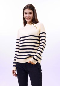 Pull rayé crème et marine avec texture côtelée, accents de boutons en or sur l'épaule et manches longues. Associé à un jeans foncé.