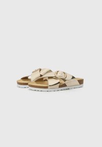 Gold metallic slide sandals with crisscross straps, a buckle detail, and a beige cork footbed atop a white textured sole.