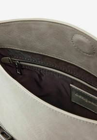 Sisätilat harmaassa keinonahkabagissä, jossa on sileä pinta, tumma verhoilu, vetoketjullinen tasku ja näkyvä brändilappu. Pyöreä sulkumechanismi.