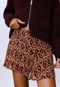 Shorts cortos y fluidos en un tejido beige que presentan un patrón geométrico en burdeos y beige, acabados con un dobladillo con volantes.