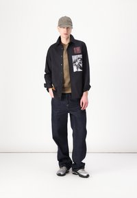 Black button-up shirt with graphic print, olive green t-shirt, dark blue jeans, gray athletic shoes, and a tan cap. Simple and casual style.