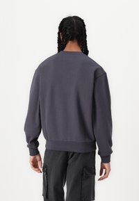 Grå sweatshirt med långa ärmar, avslappnad passform, ribbade ärmslut och nederkant, tillverkad av mjukt material. Bakifrån visas minimal detaljering och en slät textur.