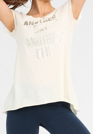 Kobieta w jasnobeżowej koszulce z napisem „ANOTHER DAY ANOTHER PLIE” i granatowych legginsach, z rękami uniesionymi nad głową.