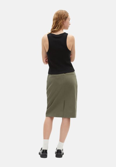 Zwarte mouwloze top en olijfgroene rok tot de knie. De rok heeft een achterkantsplit en subtiele zakken. Model draagt zwarte schoenen en witte sokken.