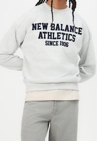 Grå sweatshirt med marinblå text som säger "NEW BALANCE ATHLETICS SINCE 1906." Har en rund hals och avslappnad passform. Bärs över en ljusfärgad skjorta.