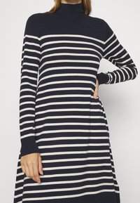 Nainen, jolla on päällään tummansininen ja valkoinen vaakasuorajuovainen pitkähihainen mekko, jossa on korkea kaulus ja ribbaustekniikalla tehdyt hihansuut, poseeraa yksi käsi lantiolla.