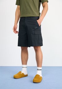 Muž oblečený v černých Nike cargo šortkách, olivově zeleném tričku, bílých ponožkách s černými pruhy a hořčicově žlutých proplétacích botách, stojící na modré podlaze.