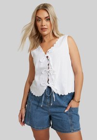 Top sin mangas de algodón blanco con dobladillo festoneado y detalle de cordones en el frente; combinado con pantalones cortos de mezclilla azul con bolsillos laterales. Textura suave.