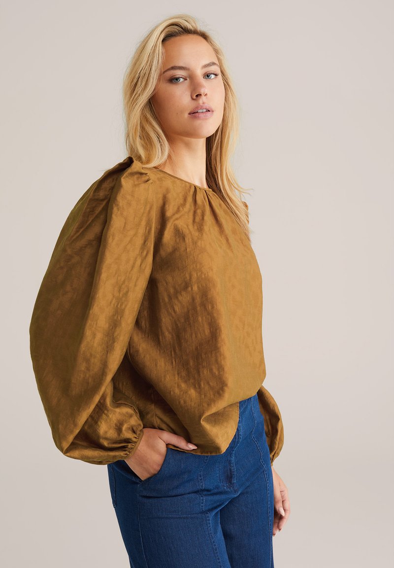 Braune, strukturierte Bluse mit Rundhalsausschnitt, Ballonärmeln und gerafften Details. Getragen mit blauen, taillenhohen Jeans.