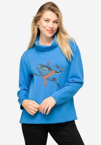 Sudadera azul claro con cuello de barco, que presenta un gráfico de una ballena con una bufanda roja. Textura suave con mangas largas y un ajuste relajado.