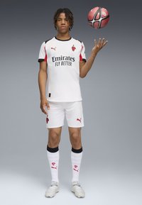 Maillot de soccer blanc avec des accents et un logo rouges, associé à un short et des chaussettes blanches montantes. Le joueur équilibre un ballon de soccer rouge et noir.