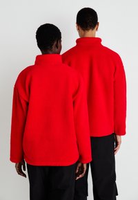 Giacche in pile rosse con colli alti e maniche lunghe, caratterizzate da una texture morbida e una vestibilità rilassata, indossate da due persone di spalle.