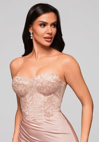 Femme aux longs cheveux noirs portant une robe corset en dentelle rose poudré sans bretelles et des boucles d'oreilles pendantes, regardant vers sa droite sur un fond gris.
