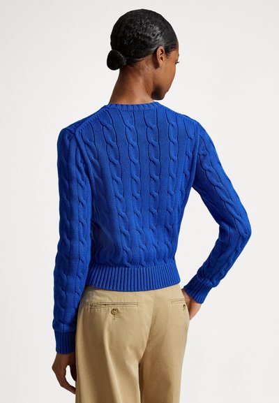 Kék, kerek nyakú, kötött mintás pulóver, texturált mintával, bordázott mandzsettákkal és szegéllyel. Világos bézs nadrág fölött viselve. Hátsó nézet.