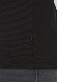 Pull en maille noir texturé avec un ourlet côtelé et une petite étiquette blanche "Blend" sur le côté. Un jean gris foncé est partiellement visible en dessous.