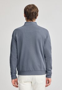 Grijze sweatshirt met een kraag en lange mouwen, met een gladde textuur en ribbels aan de manchetten, gedragen over witte broek, gezien vanuit de achterkant.