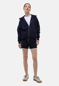 Sweat à zip marine avec cordons, associé à un short marine à bord blanc, porté avec des baskets beige et gris.