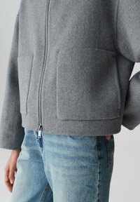 Grijze wolmix cropped jas met een rits aan de voorkant, twee grote opgestikte zakken en oversized mouwen, gecombineerd met lichtblauwe denimjeans.