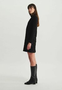 Robe mini noire pailletée à manches longues, associée à des bottes en cuir noir jusqu'aux genoux, présentant une silhouette ajustée.