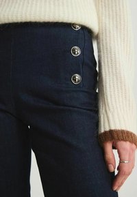 Pantalons en denim bleu marine taille haute avec des accents de boutons argentés sur le côté, assortis à un pull en tricot crème avec des poignets marron.