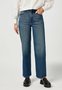 Jean bleu clair taille haute avec une coupe droite. Comprend un style classique à cinq poches et un délavage subtil. Porté avec des sabots noirs.