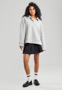 Harmaa ylisuuri collegepaita, jossa on kaulus ja etupainike, yhdistetty mustaan laskostettuun hameeseen. Mallilla on päällään mustat loafer-kengät ja raidalliset sukat.