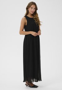 Svart plisserad maxiklänning med rund hals och ärmlös design. Geometriska plisseringar sträcker sig ned till golvet. Tillsammans med svarta klackmules.