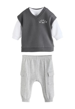 Niemowlę w koszulce z napisem "Little Man" siedzi boso na teksturowanym dywanie, patrząc na bok z otwartymi ustami obok drewnianego samochodu zabawki.