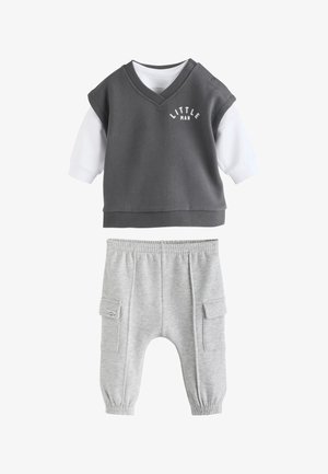 Bébé portant un t-shirt "Little Man" est assis pieds nus sur un tapis texturé, regardant sur le côté la bouche ouverte à côté d'une voiture jouet en bois.