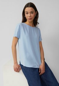 Haut bleu clair à manches courtes en tissu lisse, avec un col arrondi et un ourlet légèrement incurvé, associé à un jean en denim foncé.