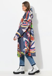 Impermeabile colorato con motivi astratti in blu, rosso e verde. Dotato di cappuccio e vestibilità ampia, abbinato a jeans e stivali argentati.