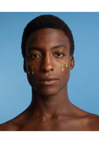 Un individuo a petto nudo con capelli corti e texturizzati guarda verso la telecamera, gocce di siero dorato su ciascuna guancia, su uno sfondo blu.