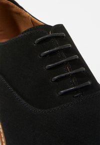 Chaussure en cuir suédé noir avec un bout fermé, un laçage et de petites perforations. Mise en valeur par un intérieur et des coutures marron contrastants.