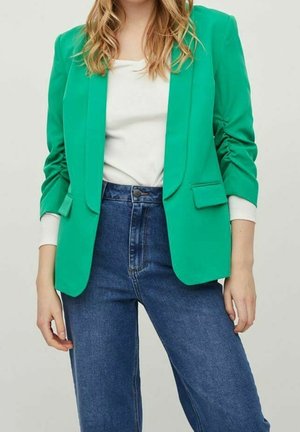 Kobieta ubrana w jasną zieloną marynarkę na białej bluzce, w połączeniu z niebieskimi dżinsami z wysokim stanem, stojąca z rękami swobodnie opuszczonymi wzdłuż ciała.