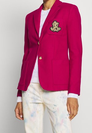 Magenta blazer met gouden geborduurd wapen en knopen, gedragen over een witte kabelgebreide trui en lichte met verfspatten bedekte broek.