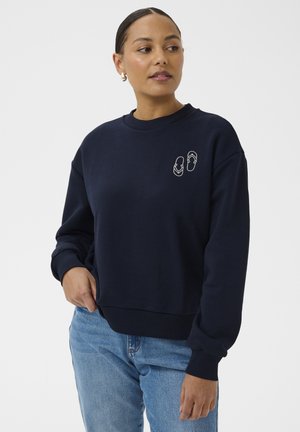 Kvinde iført en navyblå sweatshirt med et lille flip-flop-design på brystet, kombineret med lyseblå jeans, ser mod sin venstre side.