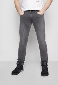 Bărbat în picioare cu mâinile în buzunare, purtând blugi gri, adidași negri înalți și un tricou alb cu logo-ul "REPLAY".