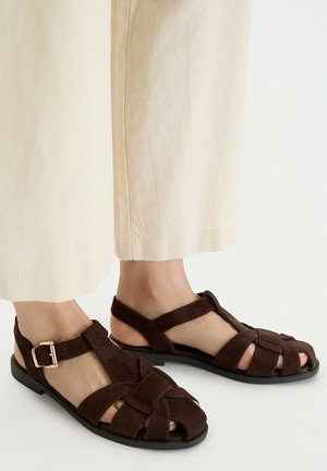 Stopy w ciemnobrązowych zamszowych sandałach zakrywających palce z paskami zapinanymi na złote klamry oraz kremowe szerokie spodnie na tle gładkiego białego tła.