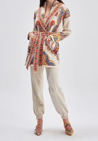 Robe beige à motifs avec des designs orange, bleu et or, taille cintrée, associée à un jogging crème et des chaussures colorées à enfiler.