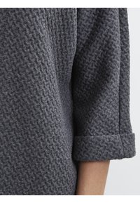 Grått texturerat tyg med ett vävt mönster, med en rullad manschett. Materialet verkar mjukt och något luddigt.