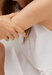 Une personne portant une robe sans manches blanche tient délicatement son bras supérieur avec sa main opposée.