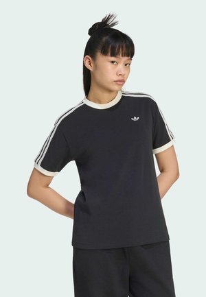 Ung person med sort hår bundet op, iført sort Adidas-trøje med hvide kanter og trestribede ærmer, stående med hænderne bag ryggen.